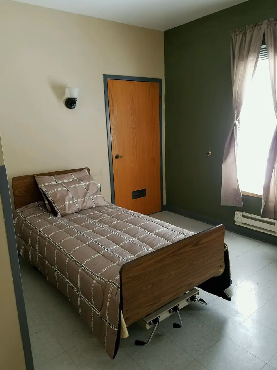Small bedroom with a single adjustable bed, wooden door, and a curtained window.