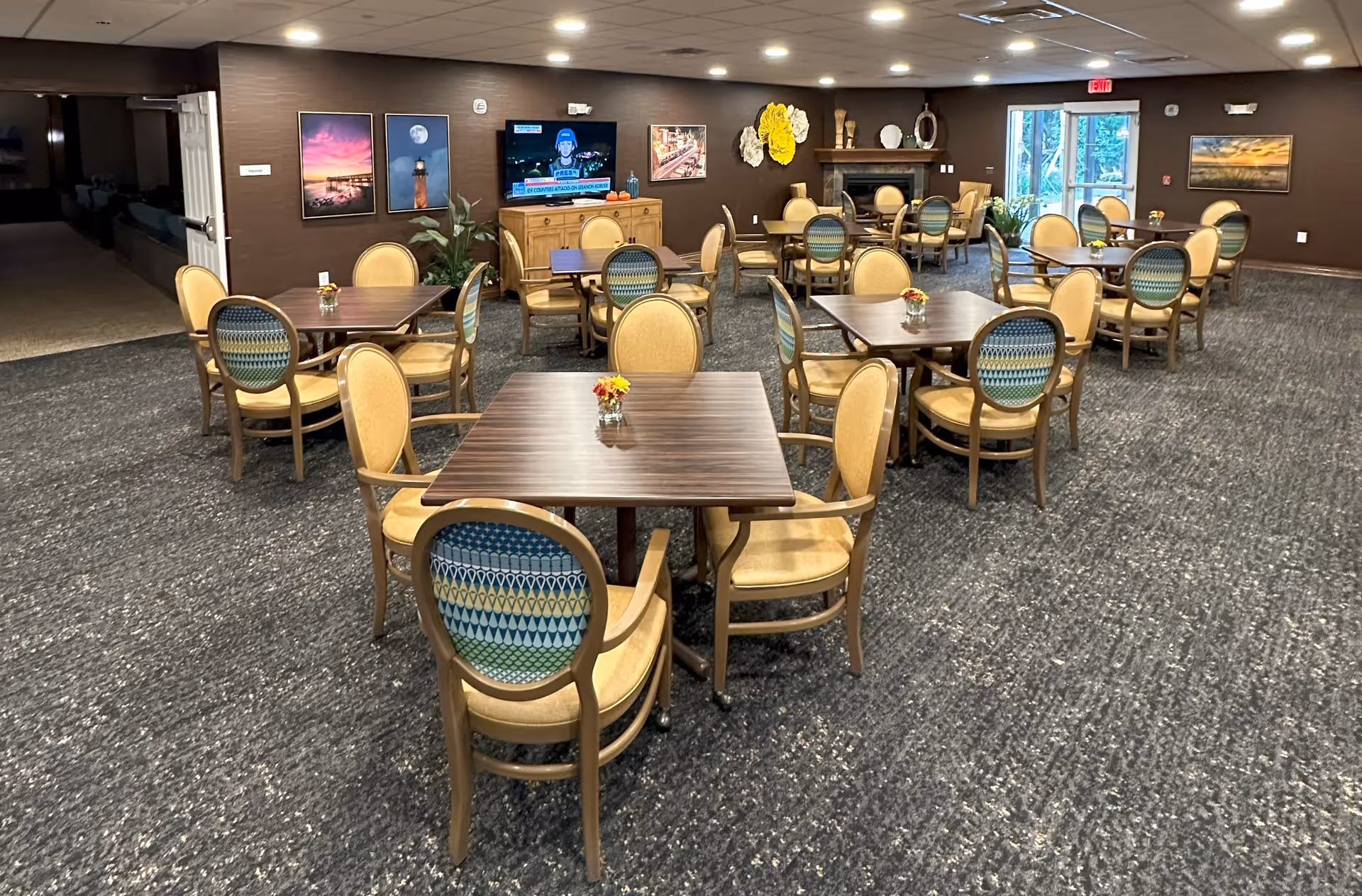 A dining room with multiple square wooden tables surrounded by cushioned chairs with patterned backs and beige seats. The room has a carpeted floor, brown walls adorned with framed pictures and decorative wall art, a TV mounted on the wall, and a fireplace. There is a glass door leading outside at the far end of the room.
