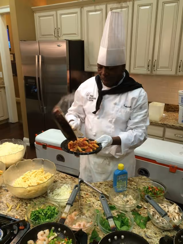 A chef wearing a white uniform and tall chef hat is preparing a plate of pasta with sauce in a kitchen. Various bowls of ingredients including pasta, chopped onions, shrimp, greens, and mushrooms are arranged on the countertop in front of him. The kitchen has cream-colored cabinets and a stainless steel refrigerator in the background.
