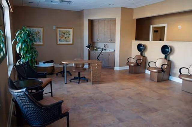 A spacious room with beige walls and tiled floor, featuring a desk with a chair, two salon hair dryer chairs, additional seating, a large plant near the window, and framed artwork on the wall.