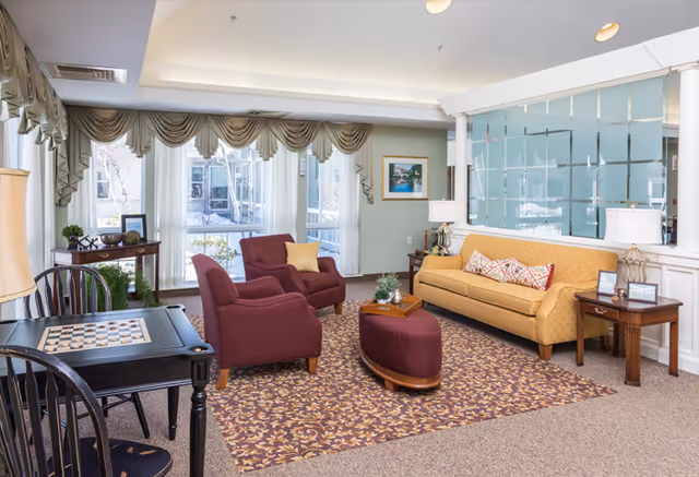 A bright and cozy living room area with a yellow sofa adorned with patterned pillows, two maroon armchairs, and a matching ottoman on a patterned rug. There is a small wooden side table with a lamp and framed photos, a chess table with chairs, large windows with draped curtains letting in natural light, and a decorative glass partition in the background.