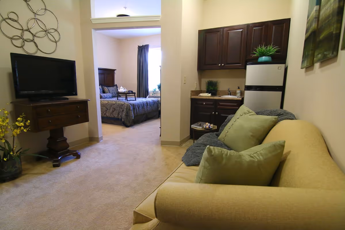 Interior view of a senior living facility apartment showing a small living area with a beige couch and green pillows, a kitchenette with dark wood cabinets and a small refrigerator, and a bedroom in the background with a bed, dark curtains, and a tray on the bed.