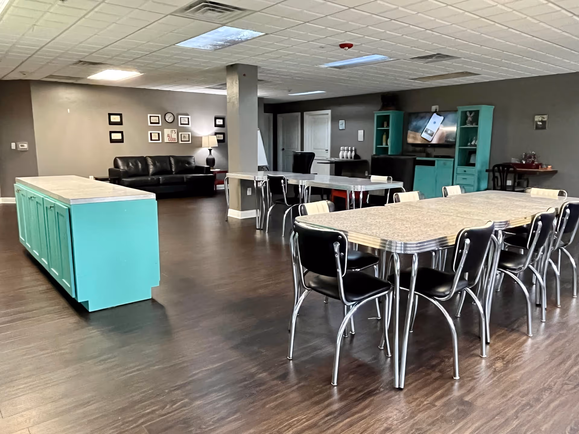 Communal dining and lounge area with retro-style tables and black chairs, teal cabinetry, and a sofa against a gray wall.