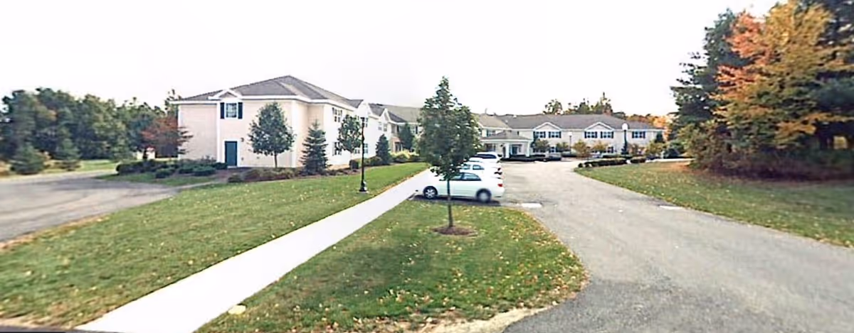 Front exterior of a beige two-story senior living building with a driveway, parked cars, sidewalk and lawns with trees.