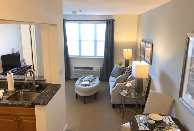 Interior view of a senior living facility apartment showing a small living room with a gray couch, patterned pillows, a round glass side table with a lamp, an oval ottoman, and a window with dark curtains. To the left, there is a kitchen area with a sink and countertop, and a dining table with place settings is partially visible on the right.