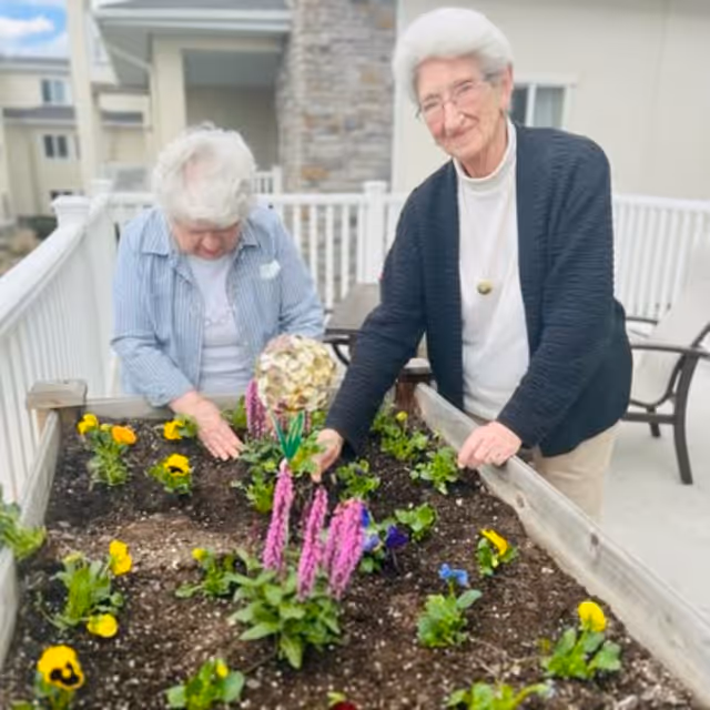 Two elderly women gardening together on a raised garden bed filled with colorful flowers on a patio with white railings and outdoor furniture in the background.
