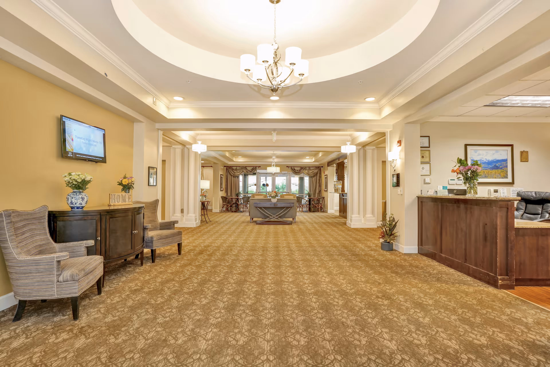 Spacious and well-lit senior living facility common area with beige patterned carpet, cream-colored walls, and a tray ceiling with a chandelier. On the left side, there are two striped armchairs and a dark wooden cabinet with flower vases and a decorative 'HOME' sign. A flat-screen TV is mounted on the wall above the cabinet. On the right side, there is a wooden reception desk with a computer and flower vase. In the background, there are more seating areas and large windows letting in natural light.