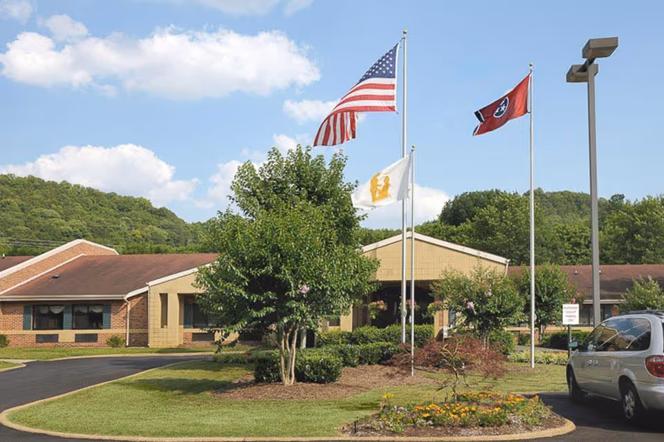 Front entrance of a single-story brick senior living facility with flagpoles, landscaped beds, and a driveway with a parked minivan.