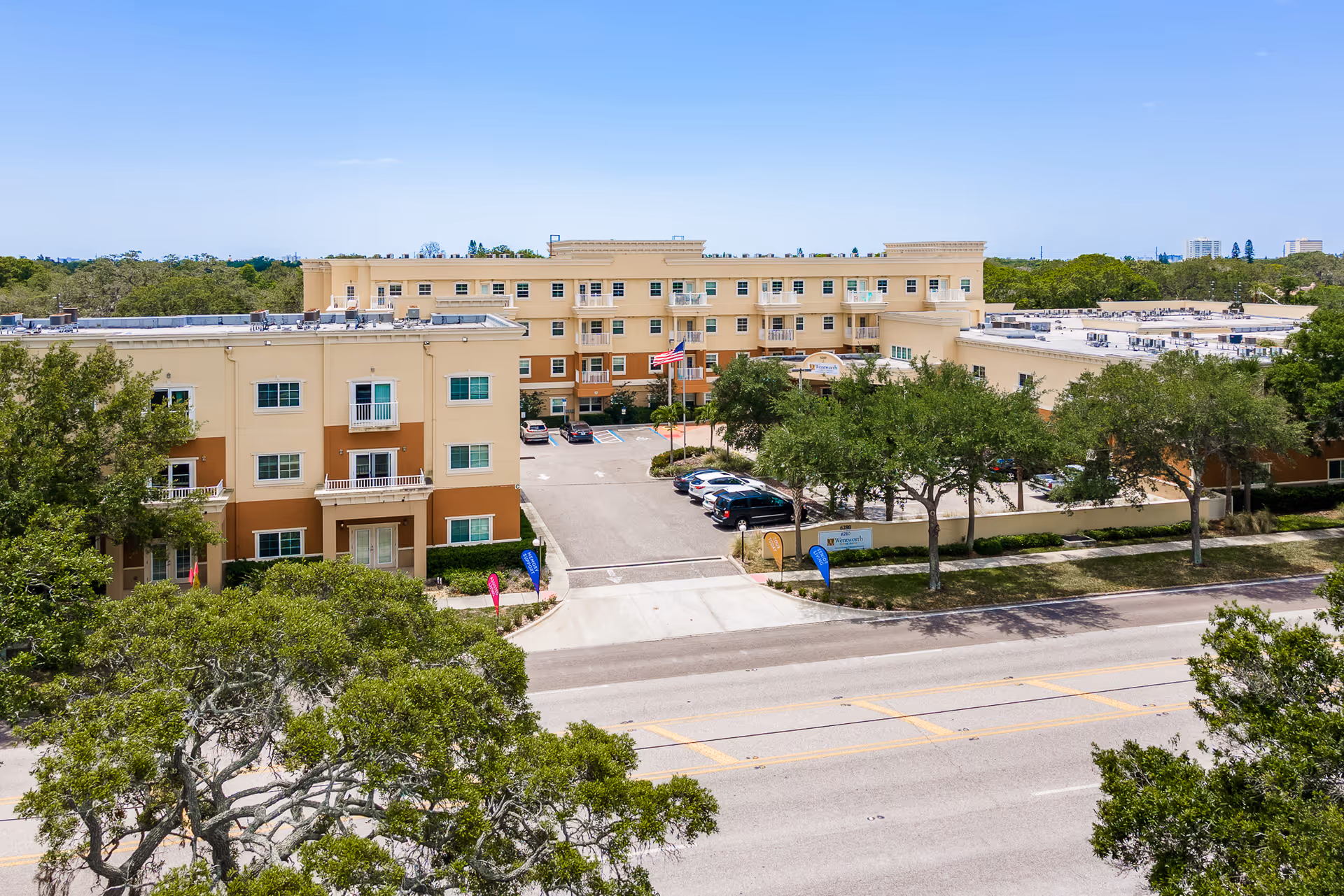 Aerial view of Wentworth Central Avenue, a multi-story senior living facility with beige and brown exterior walls, surrounded by trees and a parking lot with several cars. The building has multiple windows and balconies, and an American flag is displayed near the entrance.