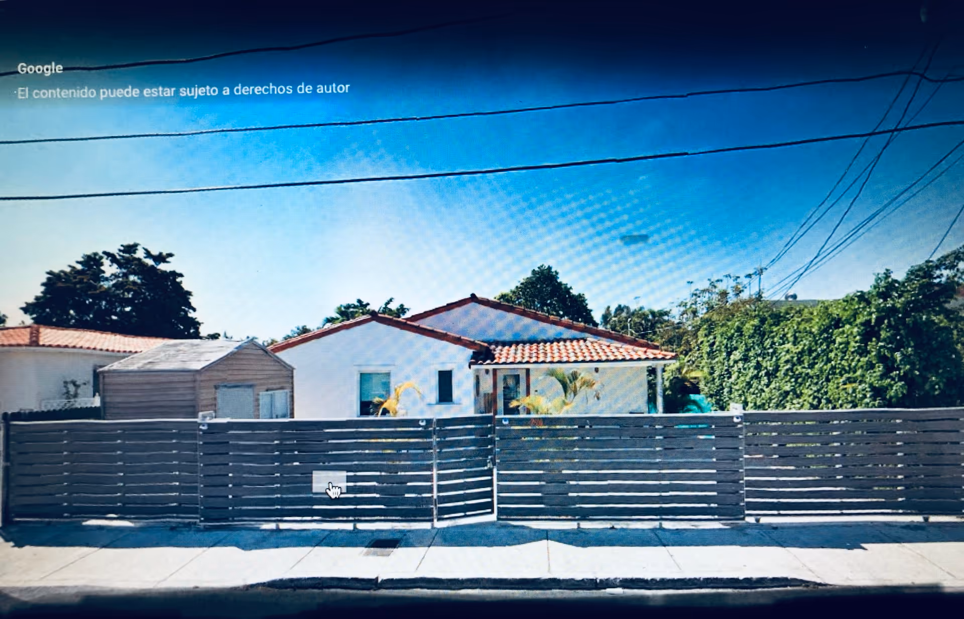 Exterior view of a single-story white building with a red-tiled roof behind a tall gray horizontal slat fence. There is a small shed to the left of the building and greenery including trees and bushes surrounding the property. Power lines are visible overhead against a clear blue sky.