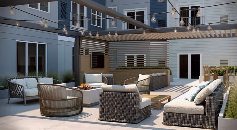 Outdoor patio area with modern wicker furniture including cushioned chairs and sofas arranged around a fire pit. Overhead string lights are hung from a pergola structure. The area is surrounded by the exterior walls of a building with windows and doors visible.