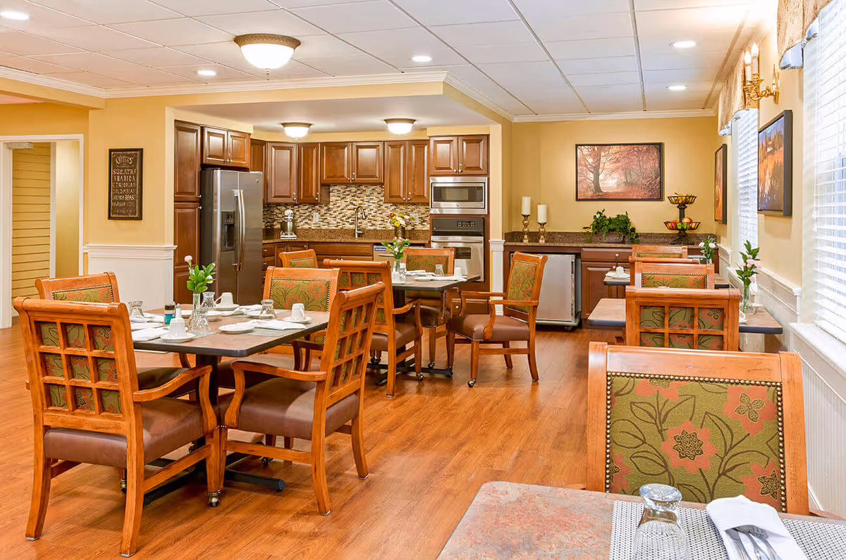 Bright dining room with wooden tables and upholstered chairs and a kitchen area in the background.