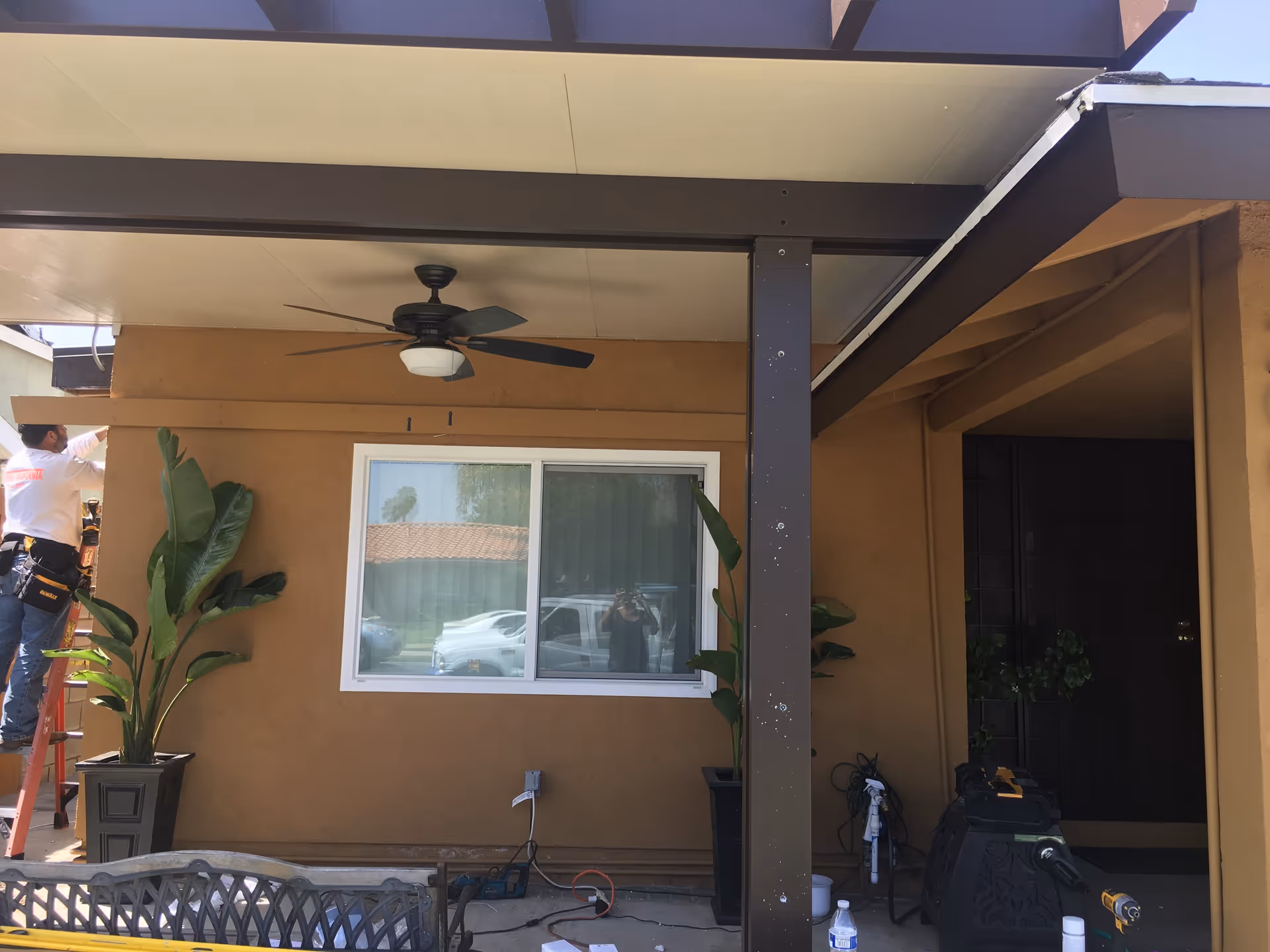 Outdoor covered patio area with a ceiling fan, two large potted plants on either side of a window, a person standing on a ladder working on the left side, and various tools and equipment on the ground.