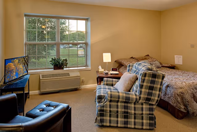 Cozy bedroom with a bed, plaid armchair, TV, nightstand lamp and a large window overlooking green lawn.