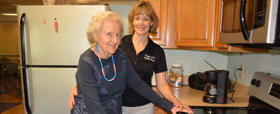An elderly woman and a caregiver standing together in a kitchen. The elderly woman is smiling and wearing a dark sweater with a beaded necklace, while the caregiver is standing behind her with a supportive arm around her waist. The kitchen has wooden cabinets, a refrigerator, a coffee maker, and a stove.