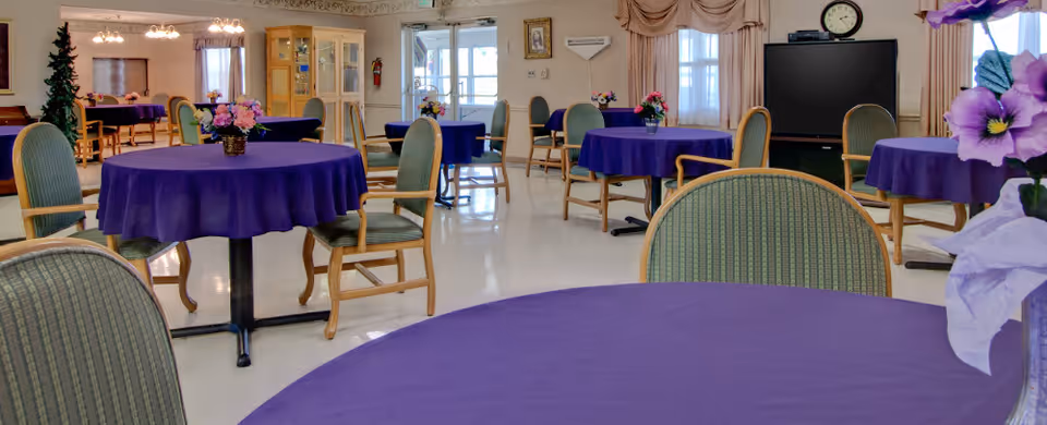 Communal dining/activity room with round tables covered in purple tablecloths, green cushioned chairs, floral centerpieces, and a TV at the back.