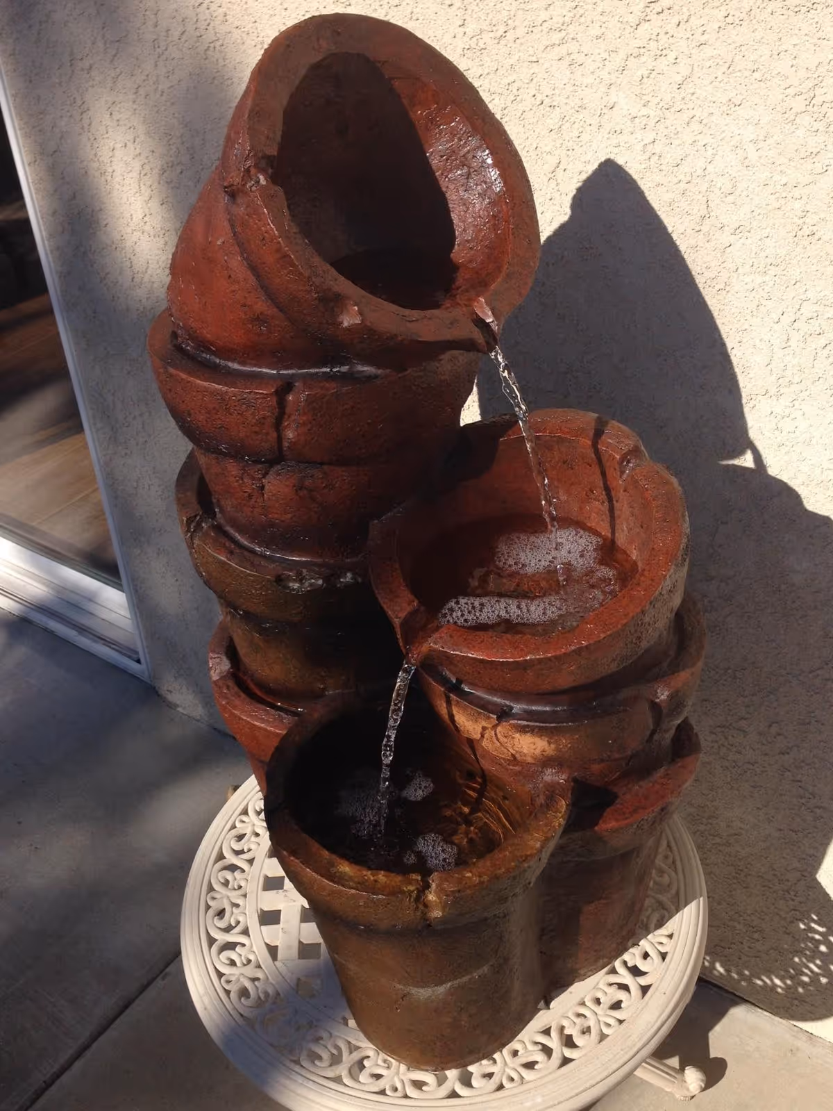 A decorative outdoor water fountain made of stacked, rustic, reddish-brown ceramic pots with water flowing from the top pot to the lower pots, placed on a white ornate metal table against a beige stucco wall.