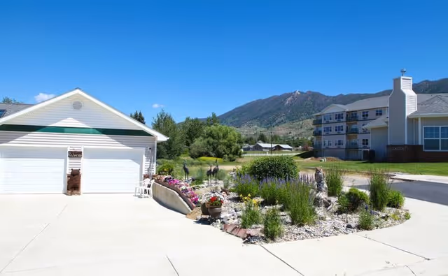 Outdoor view of a senior living facility named Big Sky on Waterford, showing a driveway with a white garage, landscaped garden with flowers and shrubs, and a multi-story building in the background with mountains under a clear blue sky.