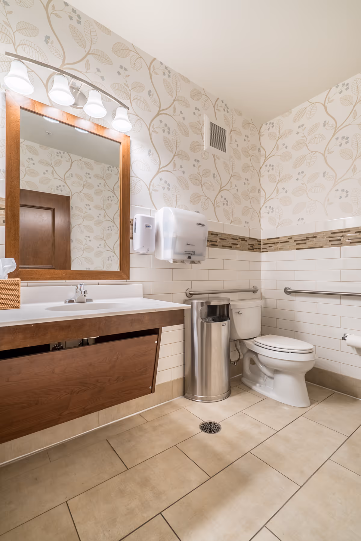 A clean and well-lit bathroom with beige tiled floor and white tiled walls with a decorative horizontal strip. The walls above the tiles have a wallpaper with a light beige leaf pattern. There is a wooden-framed mirror above a white sink with a wooden cabinet below. To the right, there is a white toilet with metal grab bars on both sides and a stainless steel trash can next to it. Above the sink, there is a wall-mounted soap dispenser and a paper towel dispenser. The bathroom has overhead lighting with four white lamps.