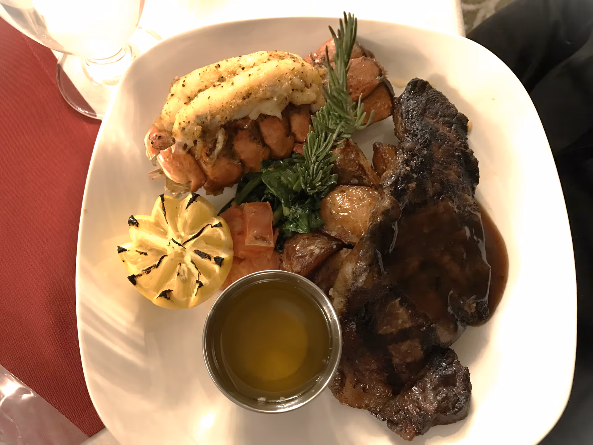 A plated meal featuring a grilled lobster tail, a grilled steak with sauce, roasted potatoes, sautéed spinach, a grilled lemon half, a small serving of diced tomatoes, and a small metal cup of melted butter, all arranged on a white plate with a red tablecloth partially visible underneath.