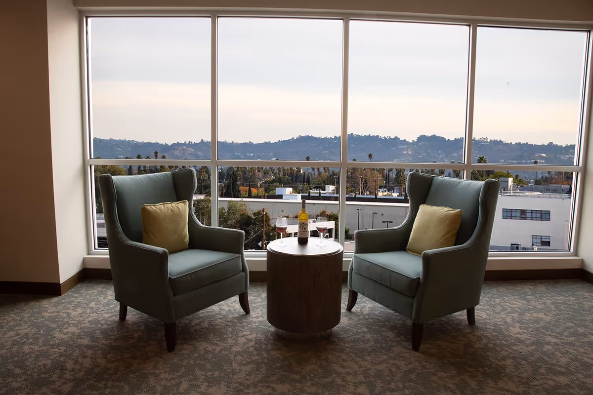 Two green armchairs with yellow pillows facing each other with a small round table between them holding a bottle of wine and two glasses, set in front of a large window showing a view of hills and buildings outside.