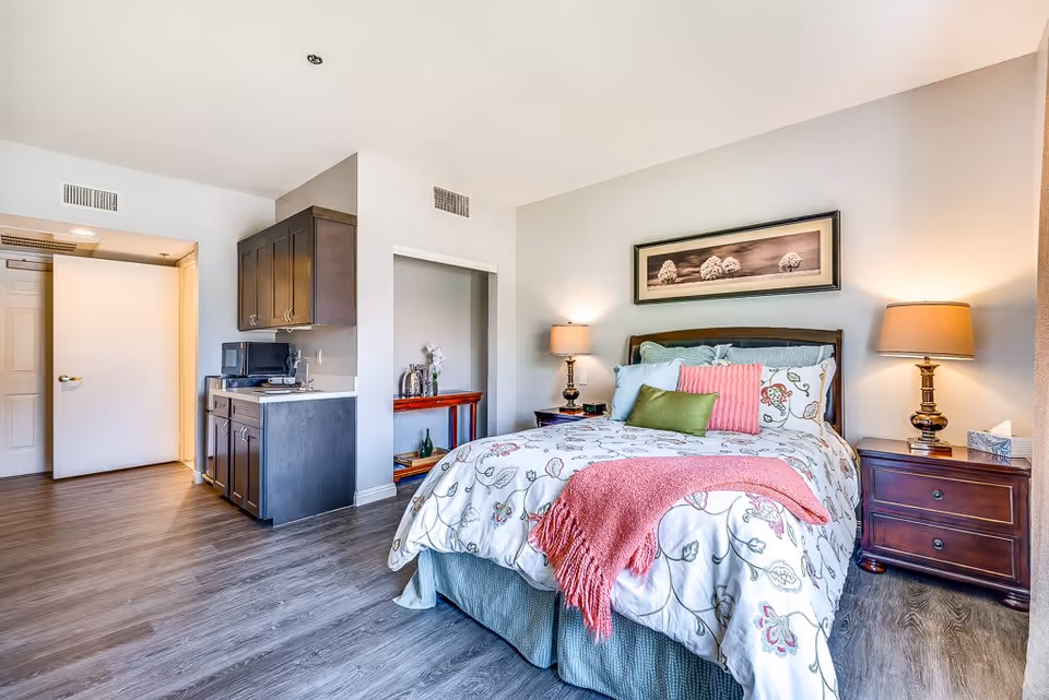 A bright and cozy bedroom with a bed featuring floral bedding, multiple pillows, and a pink throw blanket. There are two bedside tables with lamps on each side of the bed. The room has wooden flooring and light-colored walls. To the left, there is a small kitchenette area with dark cabinets, a microwave, and a sink. A door is open leading to another room or hallway.