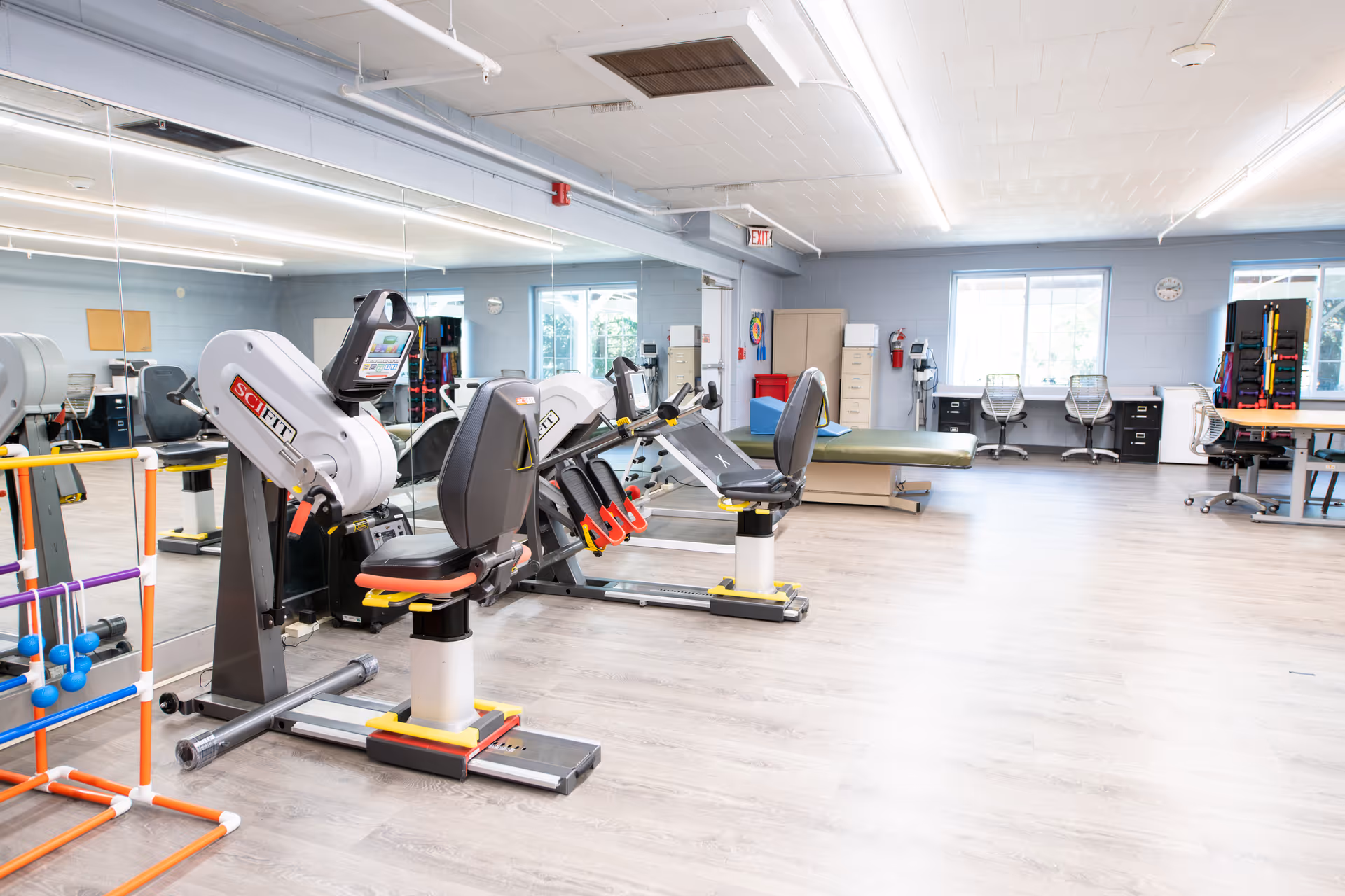 Bright rehabilitation gym with exercise machines, therapy tables, mirrors, and workstations.