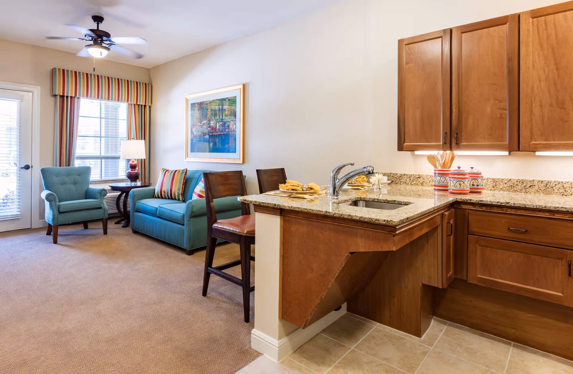 A cozy living room and kitchen area in a senior living facility. The living room features a teal armchair and matching sofa with colorful striped pillows, a side table with a lamp, and a large window with striped valances. The kitchen area has wooden cabinets, a granite countertop with a sink, and two wooden chairs at the counter.