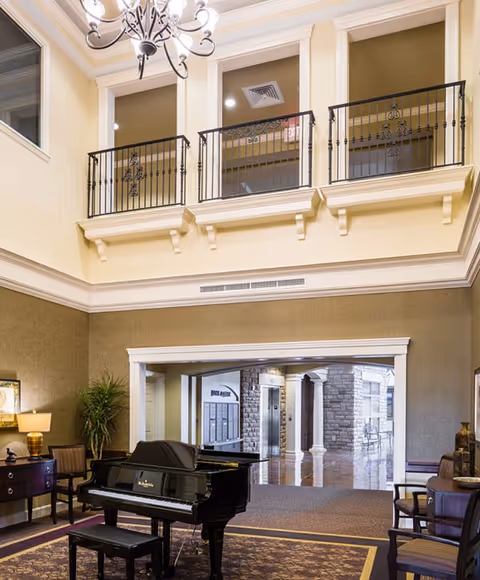 Interior view of a senior living facility lounge area featuring a black grand piano with a matching bench on a patterned rug. The room has high ceilings with a chandelier, beige walls, and a balcony with decorative black railings on the upper level. There are chairs and a side table with a lamp and plant on the left side, and an open doorway leading to a hallway with stone walls and an elevator in the background.