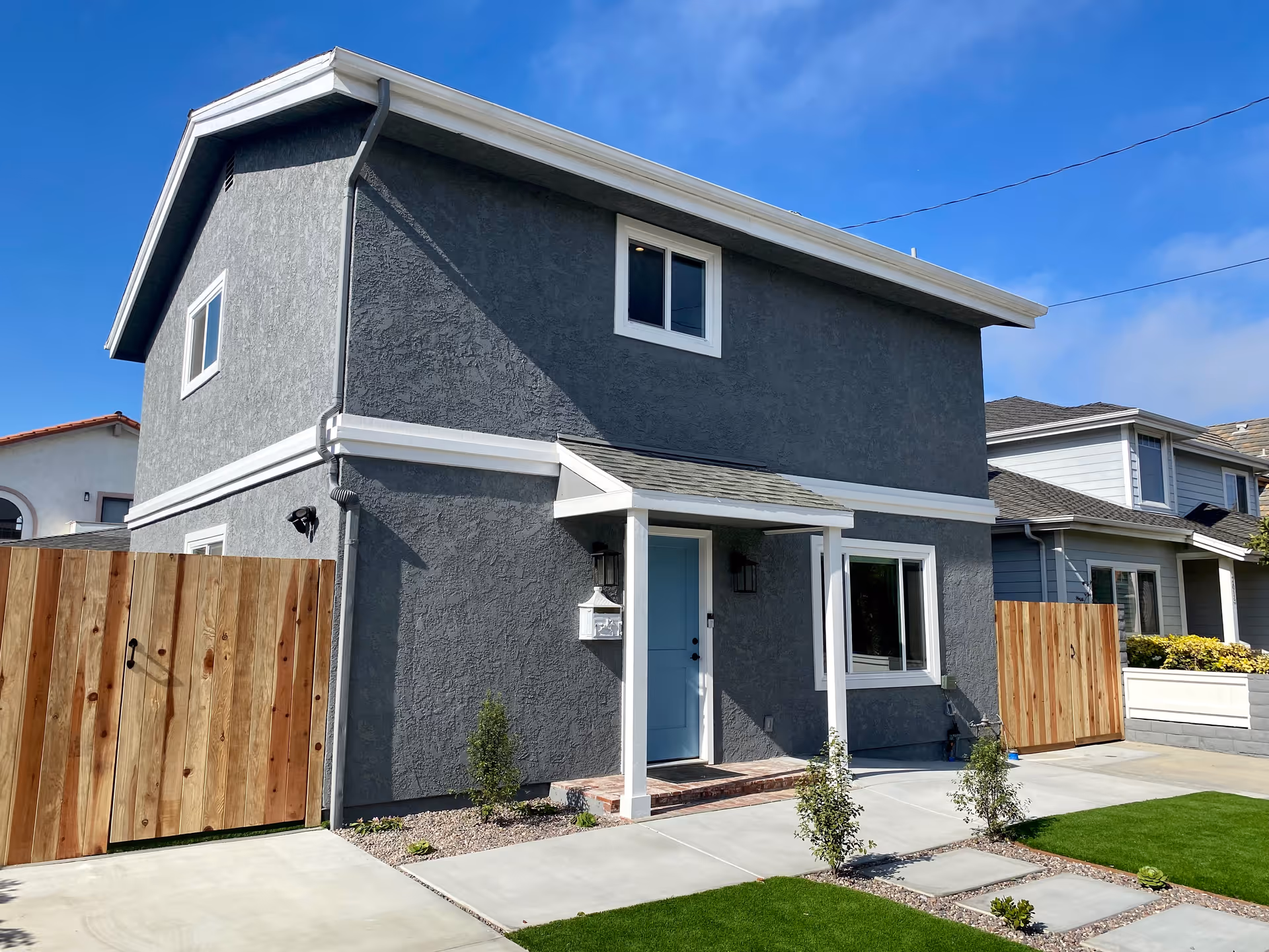 Two-story gray stucco house with a blue front door, small porch, wooden side gates and a landscaped front yard.