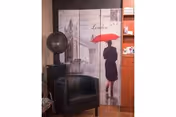 Interior corner of a room featuring a black leather armchair, a vintage-style globe on a stand, and a decorative folding screen with an image of a person holding a red umbrella walking in a rainy London street.