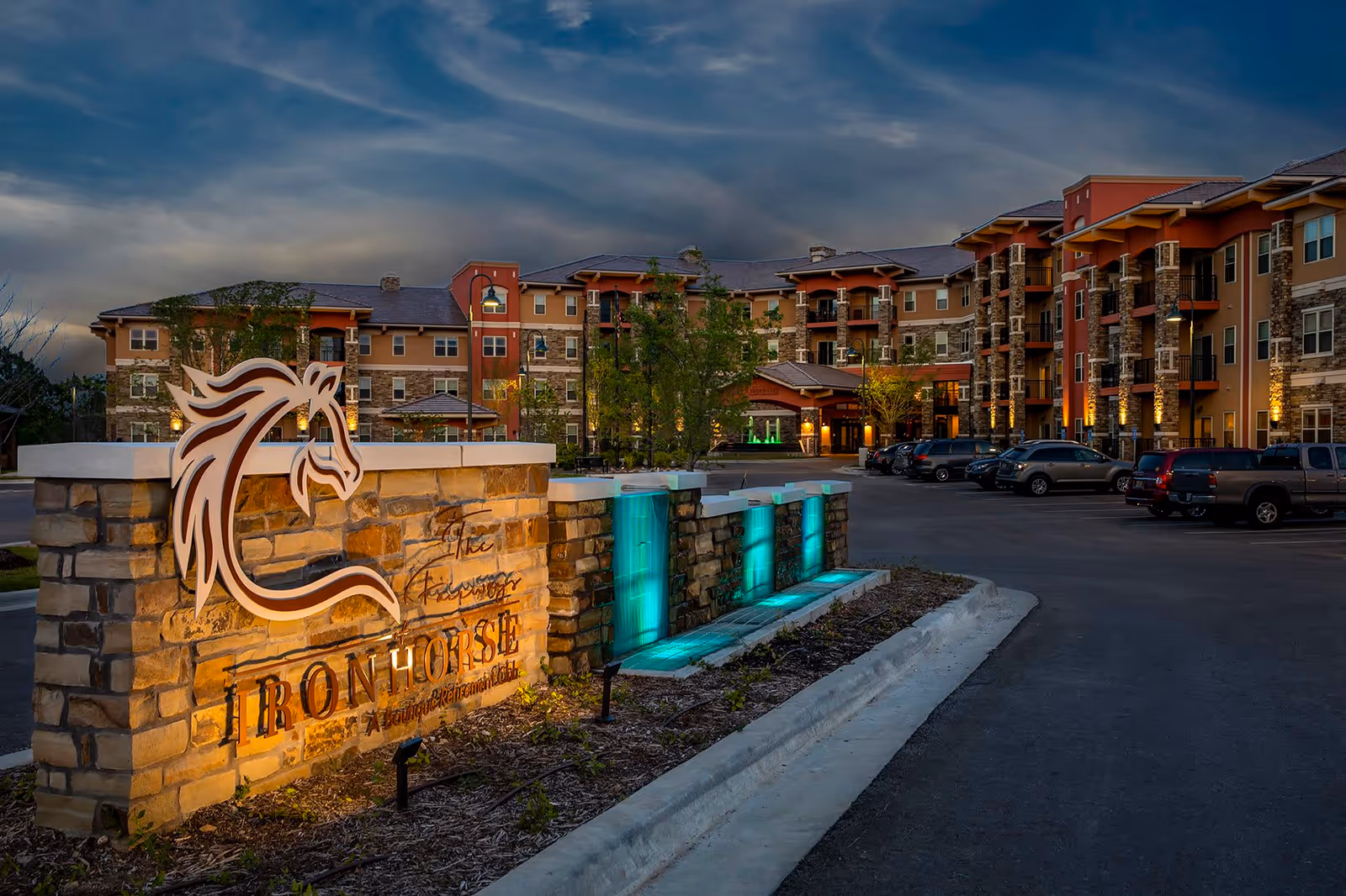 Evening view of the Fairways of Ironhorse Independent Living facility showing a large stone sign with a horse logo and the facility name illuminated by lights. Behind the sign is a multi-story residential building with balconies and warm exterior lighting. Several cars are parked in the parking lot to the right.