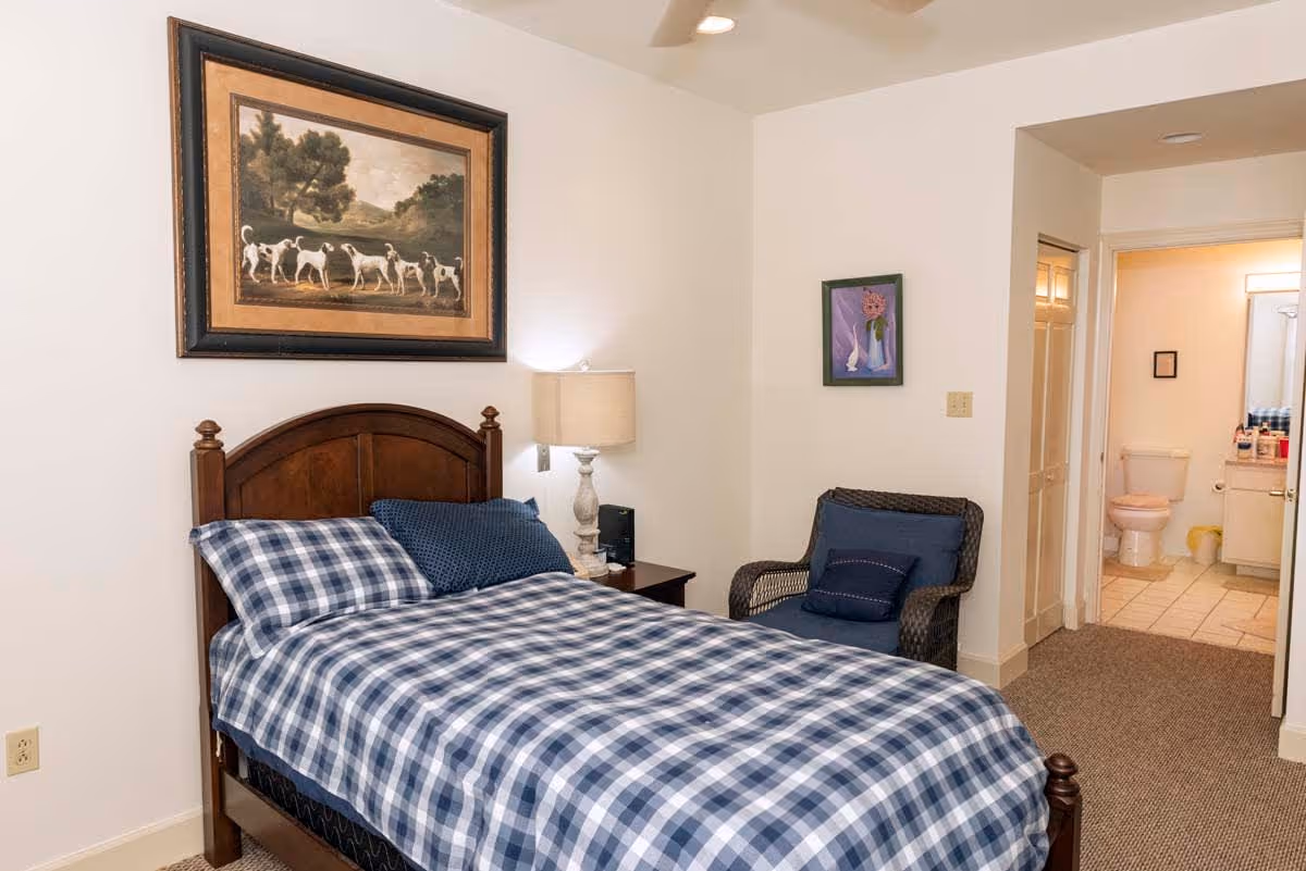 A bedroom with a wooden bed frame and a blue and white checkered bedspread. There is a nightstand with a lamp next to the bed, a wicker chair with blue cushions, and a painting of dogs hanging above the bed. In the background, an open doorway leads to a bathroom with a toilet and sink visible.