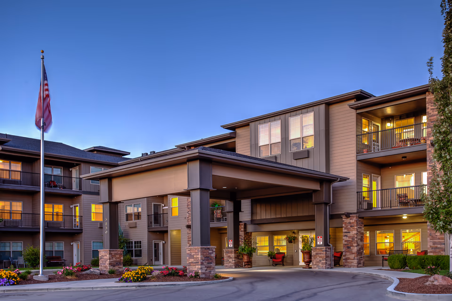 Exterior view of a three-story senior living facility building at dusk with warm lights glowing from the windows. The entrance features a covered drop-off area supported by stone pillars. An American flag is flying on a flagpole near the entrance, and there are landscaped flower beds around the driveway.