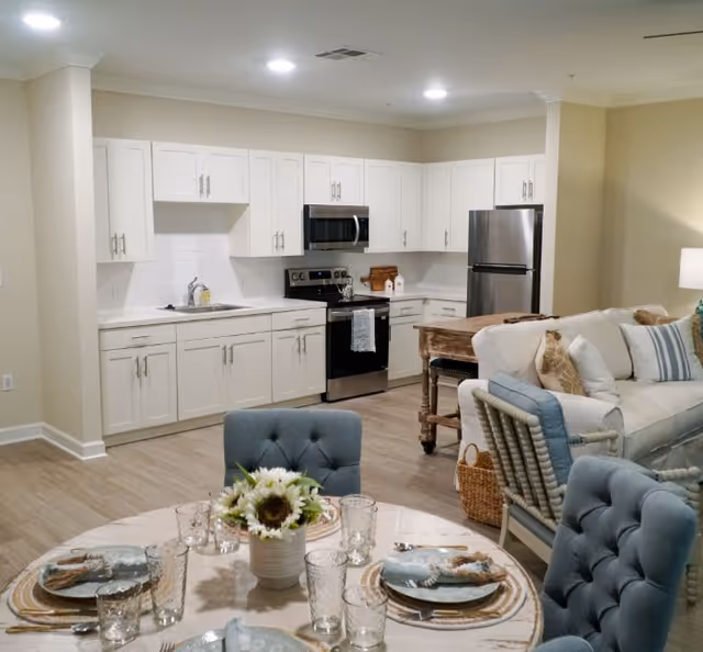 A modern open-concept kitchen and dining area with white cabinets, stainless steel appliances including a refrigerator, stove, and microwave. The dining table is set with plates, glasses, and napkins, and has a centerpiece with sunflowers. Adjacent to the kitchen is a cozy living area with a white sofa and blue cushioned chairs.