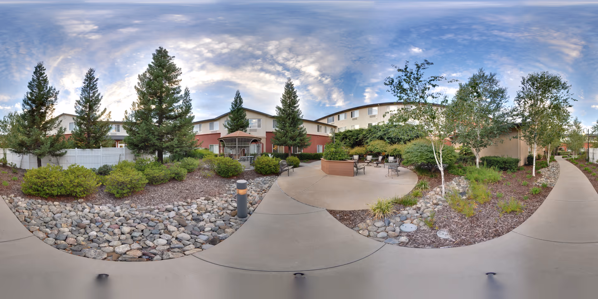 Courtyard with paved walkways, seating area, trees and landscaped rock beds in front of a two-story senior living building under a partly cloudy sky.