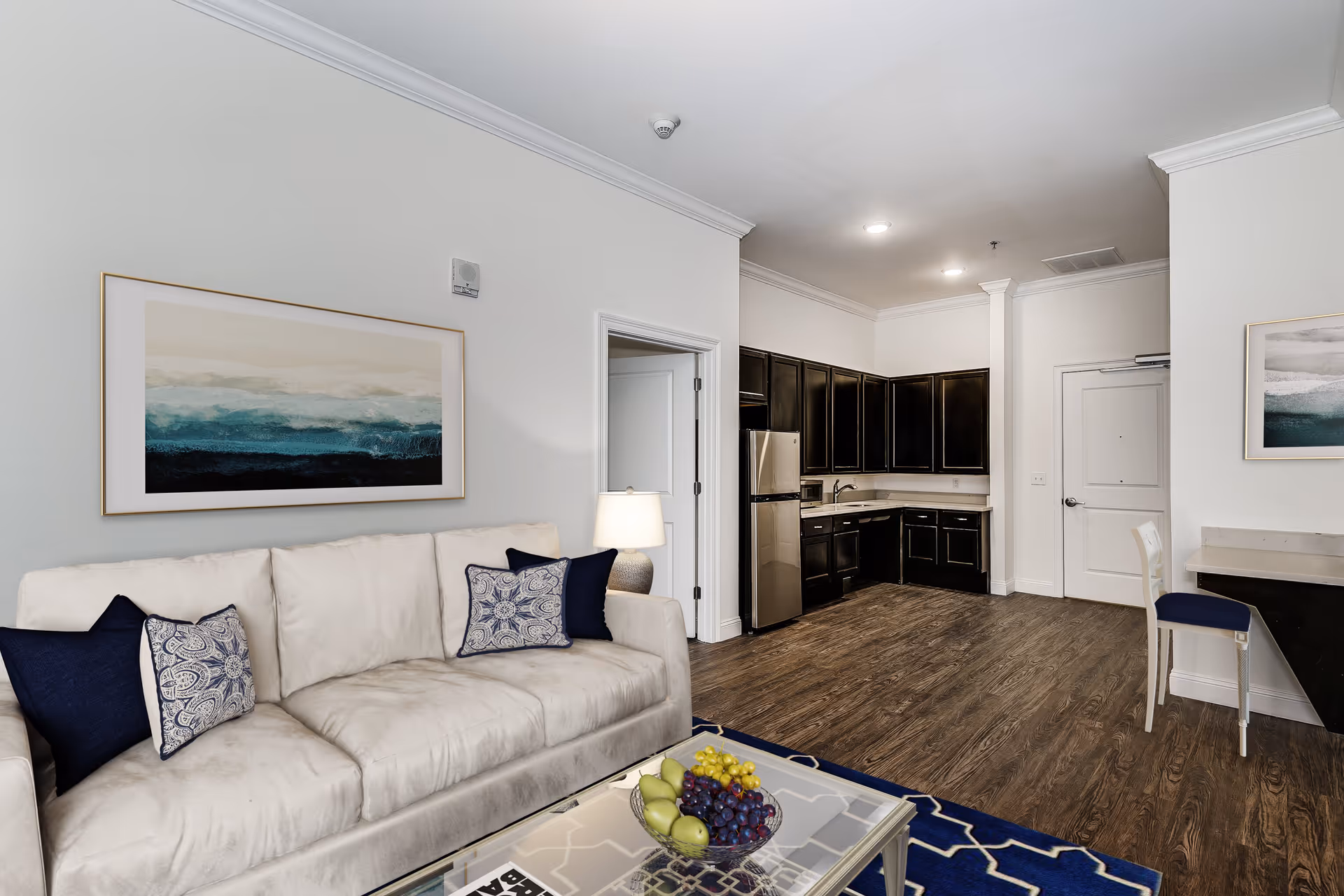 A modern living area with a beige sofa adorned with blue and patterned pillows, a glass coffee table with a bowl of fruit, and a blue patterned rug. The room opens into a kitchen with dark wood cabinets, stainless steel refrigerator, and a sink. The walls are white with two abstract paintings featuring blue and beige tones. There is a white chair at a small counter near the entrance door.