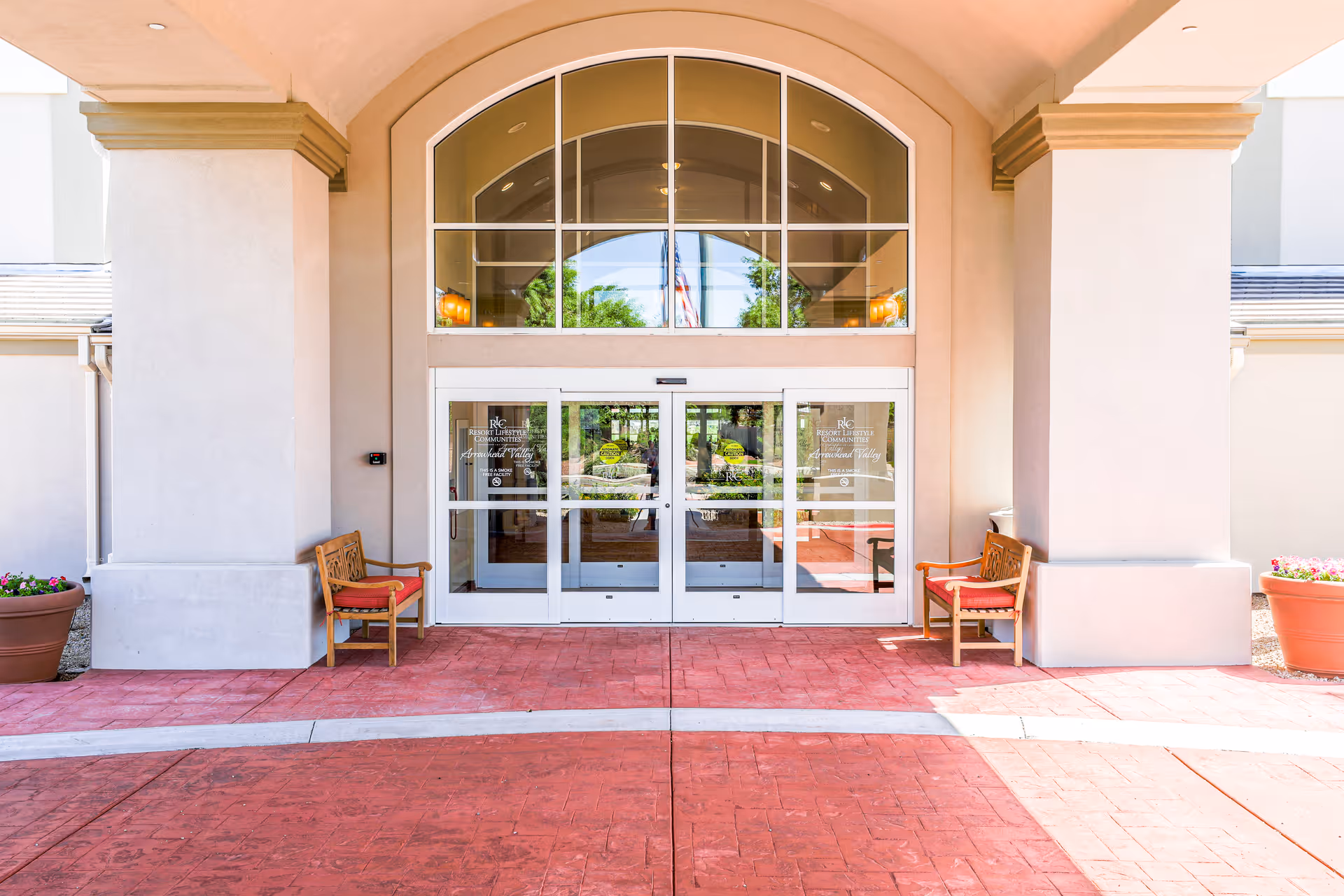 Entrance to Arrowhead Valley Retirement Resort featuring double glass doors with white frames, two wooden benches with red cushions on either side, large potted plants, and an arched window above the doors.
