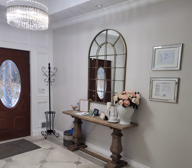 Foyer with a decorative oval-glass wooden front door, a console table topped with a large arched mirror and floral arrangement, and framed certificates on the wall.