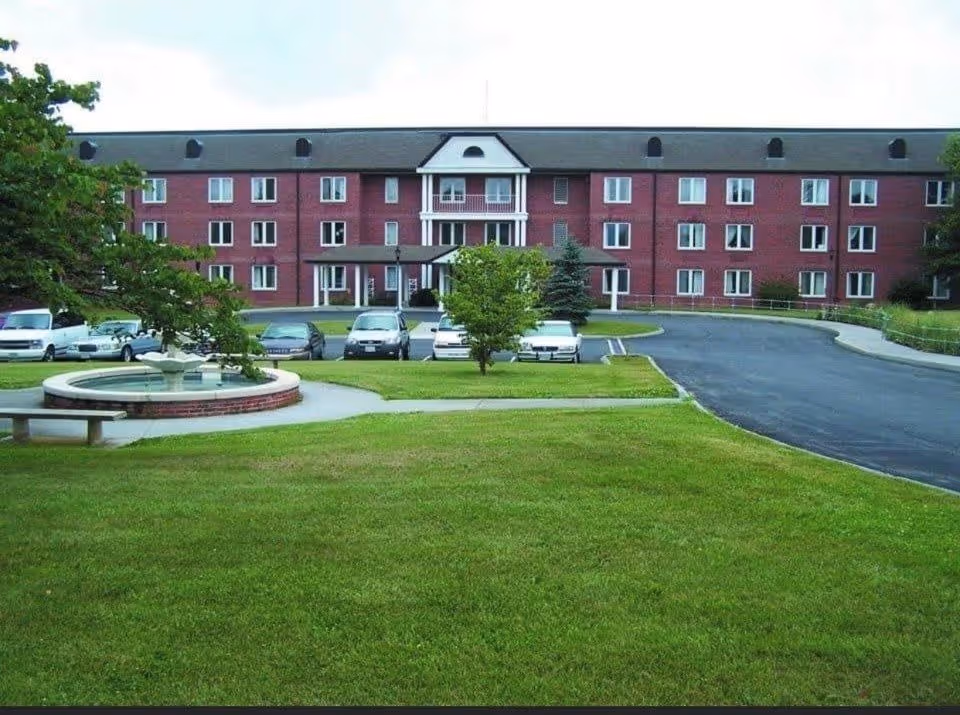Front exterior view of a three-story brick building with multiple windows, a central entrance with a small covered porch, a circular fountain surrounded by a brick border and benches, a green lawn, several trees, and a parking area with cars.
