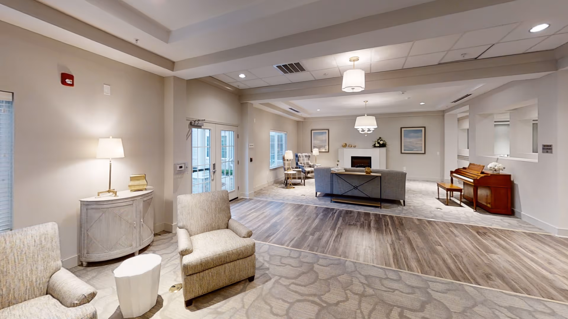 A spacious and well-lit living room area with beige walls and a mix of carpet and wood flooring. The room features comfortable armchairs, a sofa, a small table with a lamp, a piano with a bench, and framed artwork on the walls. There are windows and glass doors allowing natural light to enter the space.