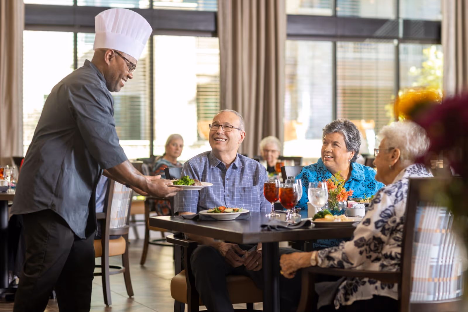 A chef serves a plate to smiling elderly residents seated at a dining table in a bright dining room.