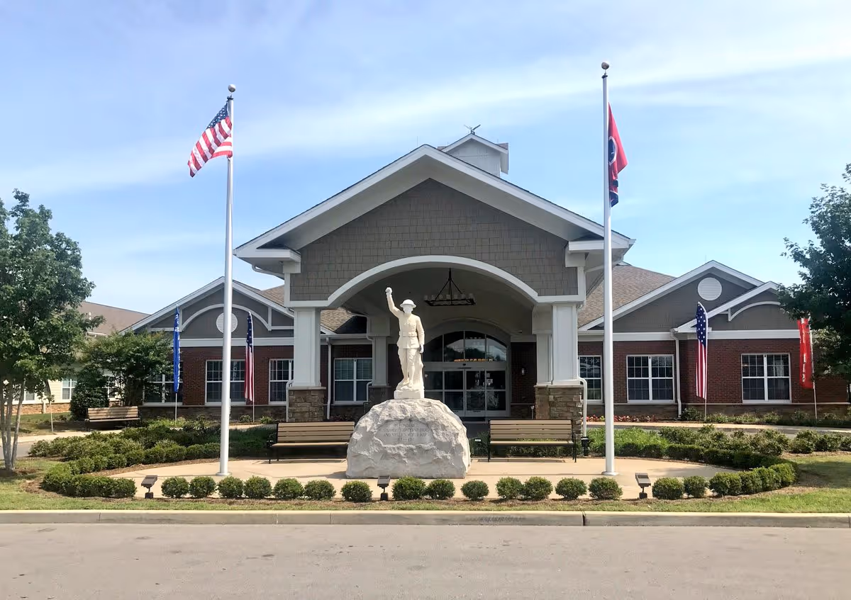 Front exterior view of the Tennessee State Veterans' Home building with a covered entrance, a statue of a soldier on a rock in the center, two flagpoles with the American flag and Tennessee state flag, benches, and landscaped greenery.