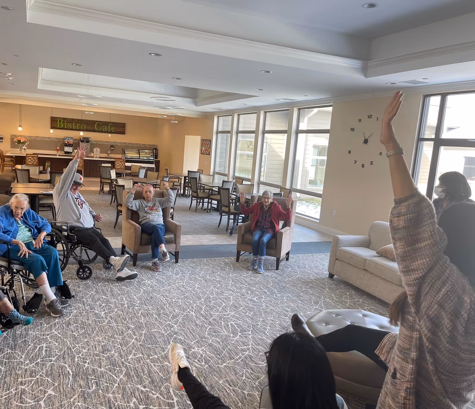 A group of elderly people and two caregivers participating in a seated exercise or activity in a spacious, well-lit room with large windows. The room has a carpeted floor, several chairs, and tables in the background, and a sign that reads 'Bistro Cafe' above a counter area.