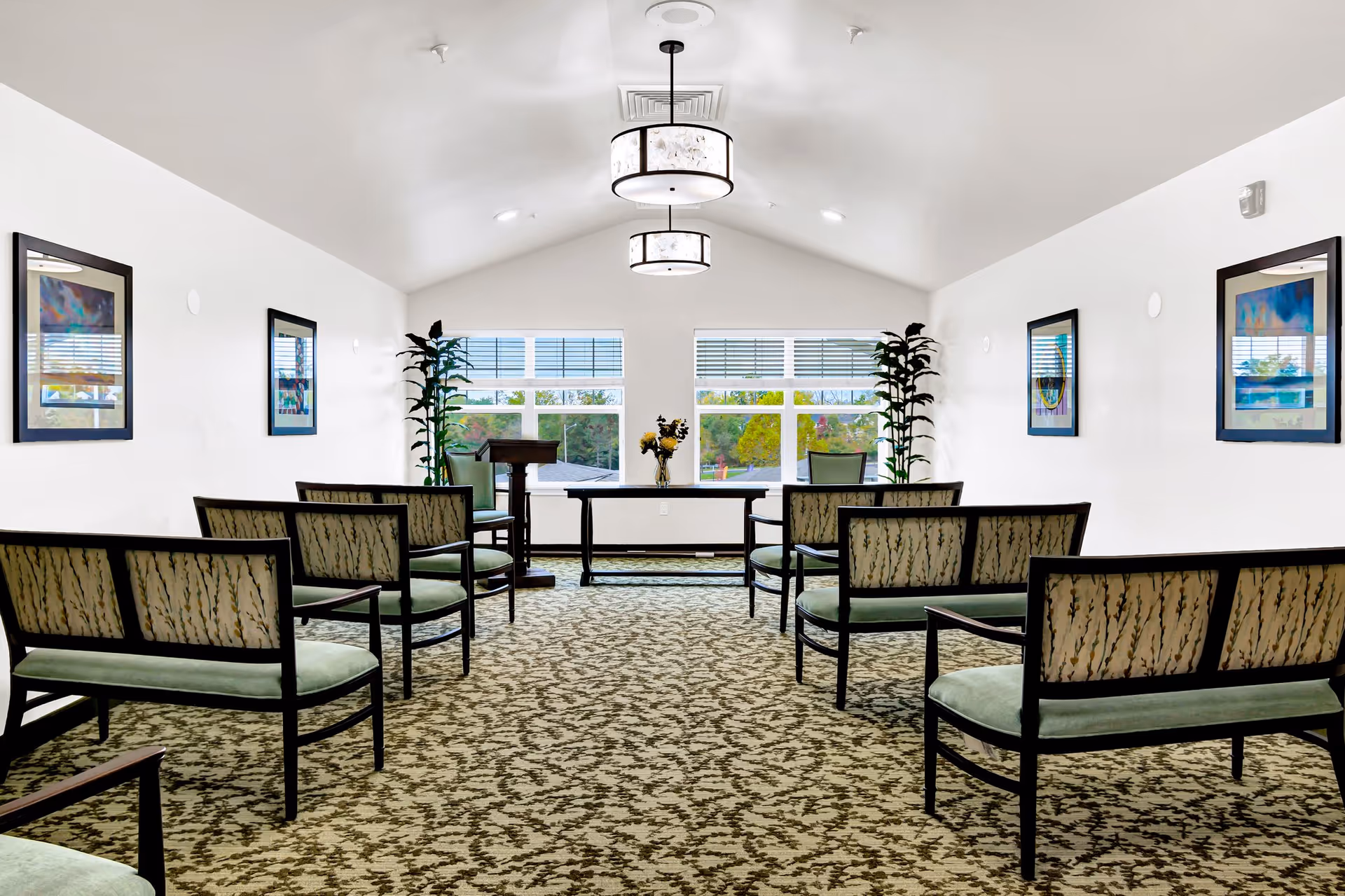 A bright, well-lit room with patterned carpet and rows of green cushioned chairs facing a table and podium near two large windows. The walls are white with framed artwork and tall plants near the windows. Two ceiling lights hang from the vaulted ceiling.