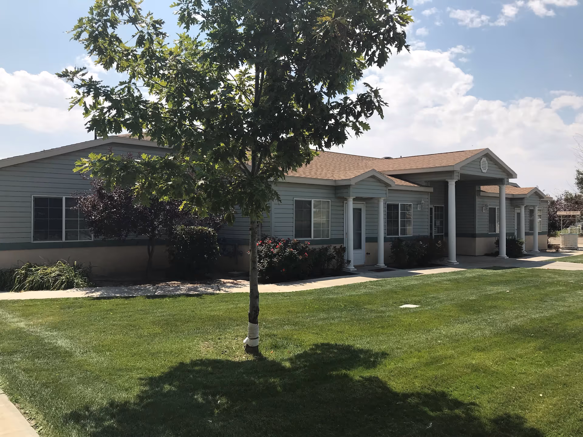 Exterior view of a single-story senior living facility building with light gray siding, white columns at the entrance, several windows, and a well-maintained green lawn with a tree in the foreground under a partly cloudy sky.