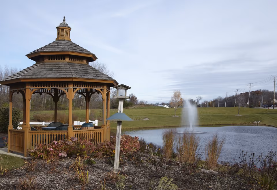 A wooden gazebo beside a pond with a fountain and a birdhouse in a grassy landscape.