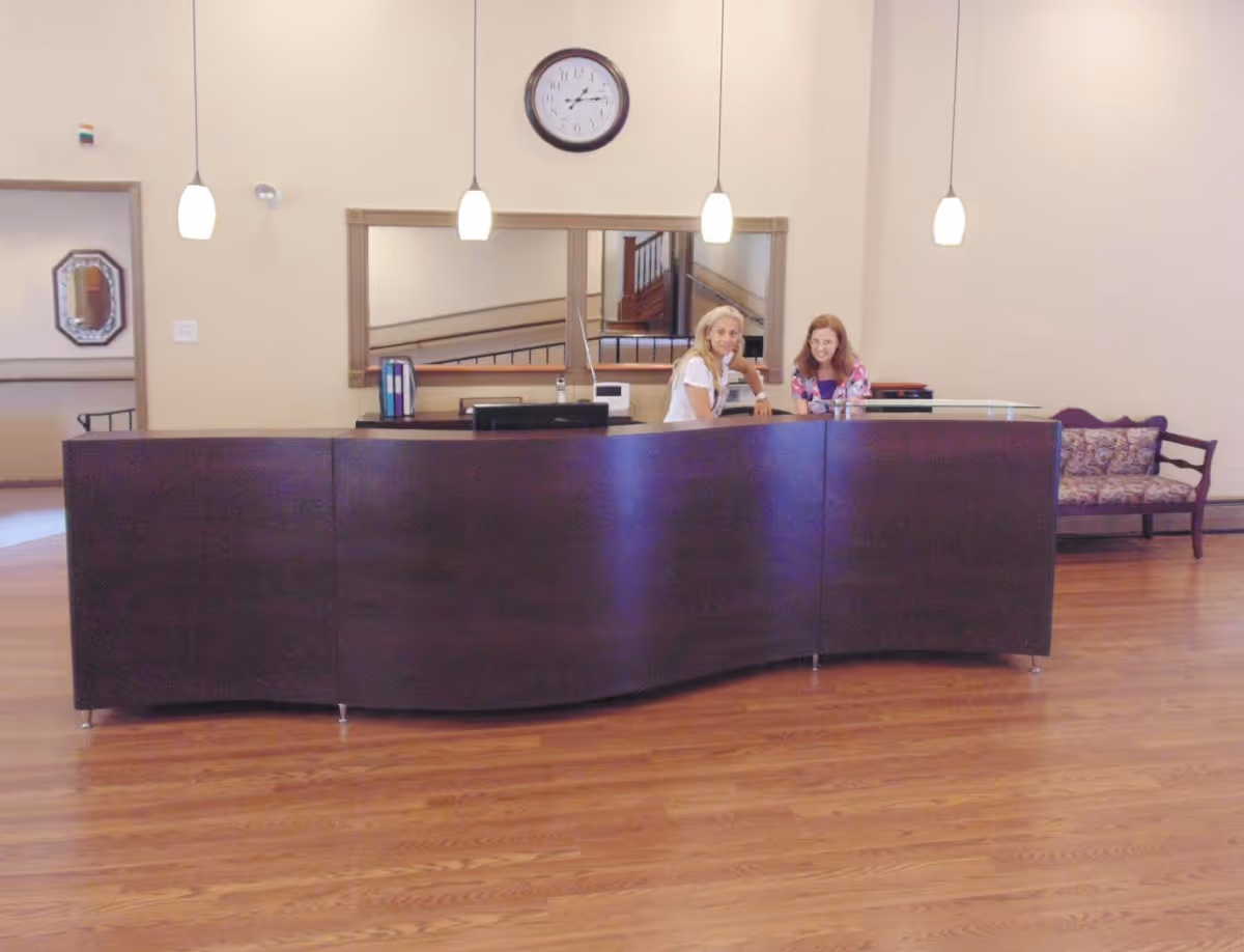 Reception area with a curved dark wood desk, two women sitting behind it, a wall clock above, pendant lights hanging from the ceiling, a large mirror on the wall, and a wooden bench with patterned cushions to the right.