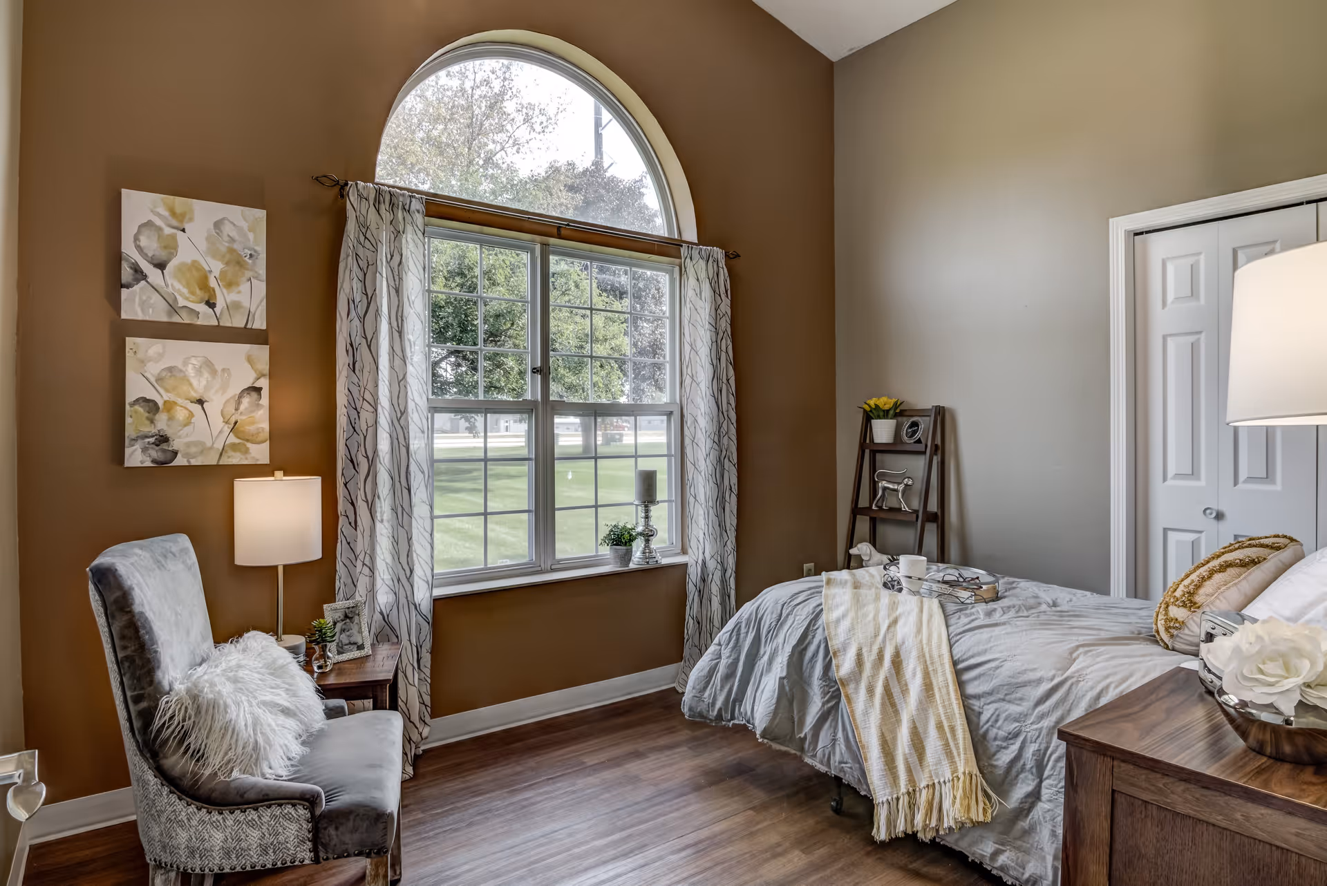 A cozy bedroom with a large arched window letting in natural light. The room features a bed with gray bedding and a yellow striped throw blanket, a wooden nightstand with a lamp and decorative items, a gray upholstered chair with a fluffy white pillow, and a small ladder shelf with plants and decor. The walls are painted in warm tones and the floor is wooden.