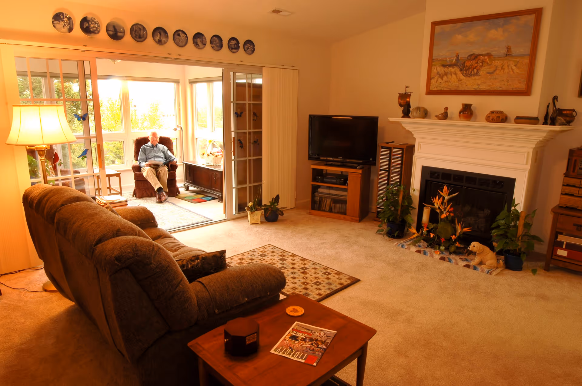 A cozy living room with a brown sofa, a wooden side table with a magazine and a candle, a white fireplace decorated with plants and small sculptures, and a flat-screen TV on a wooden stand. In the background, an elderly man is sitting in a recliner chair in a sunlit enclosed porch area with large windows and sliding glass doors. The walls are adorned with decorative plates and a framed painting above the fireplace.